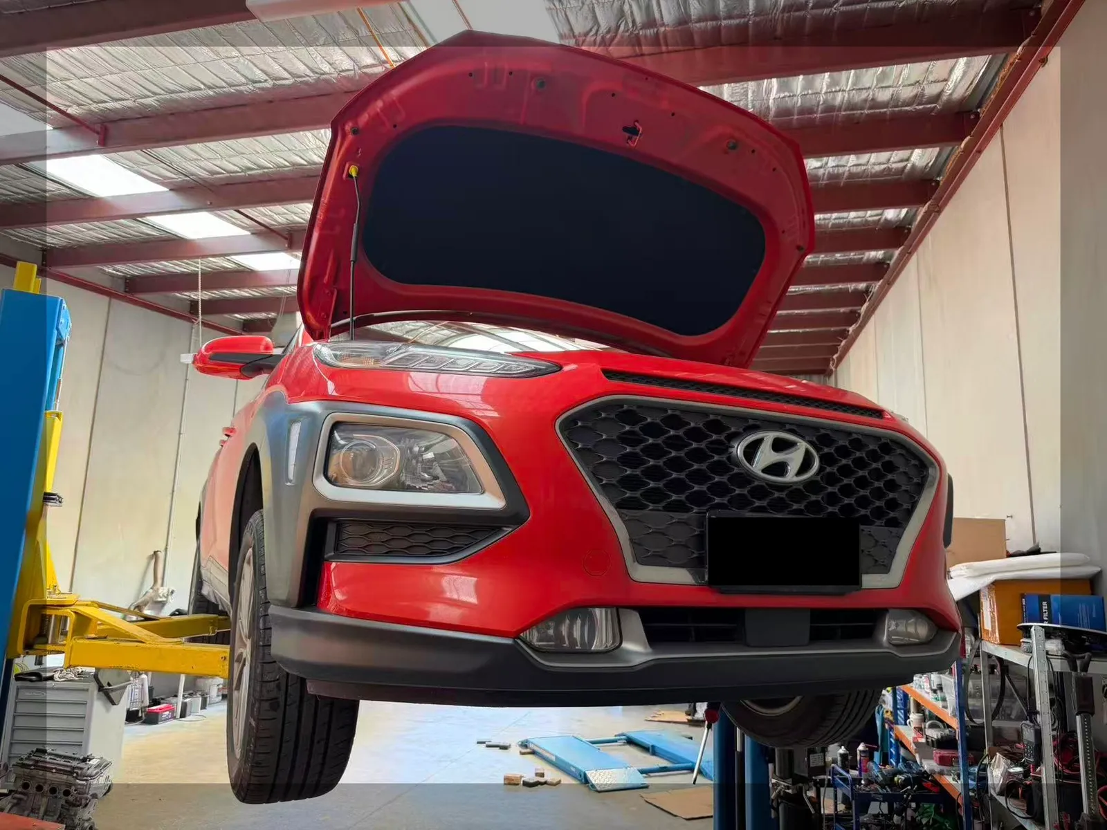 Red Hyundai Kona raised on a two-post hoist with the bonnet open at C1 Automotive in Oakleigh South, Melbourne, with the number plate redacted