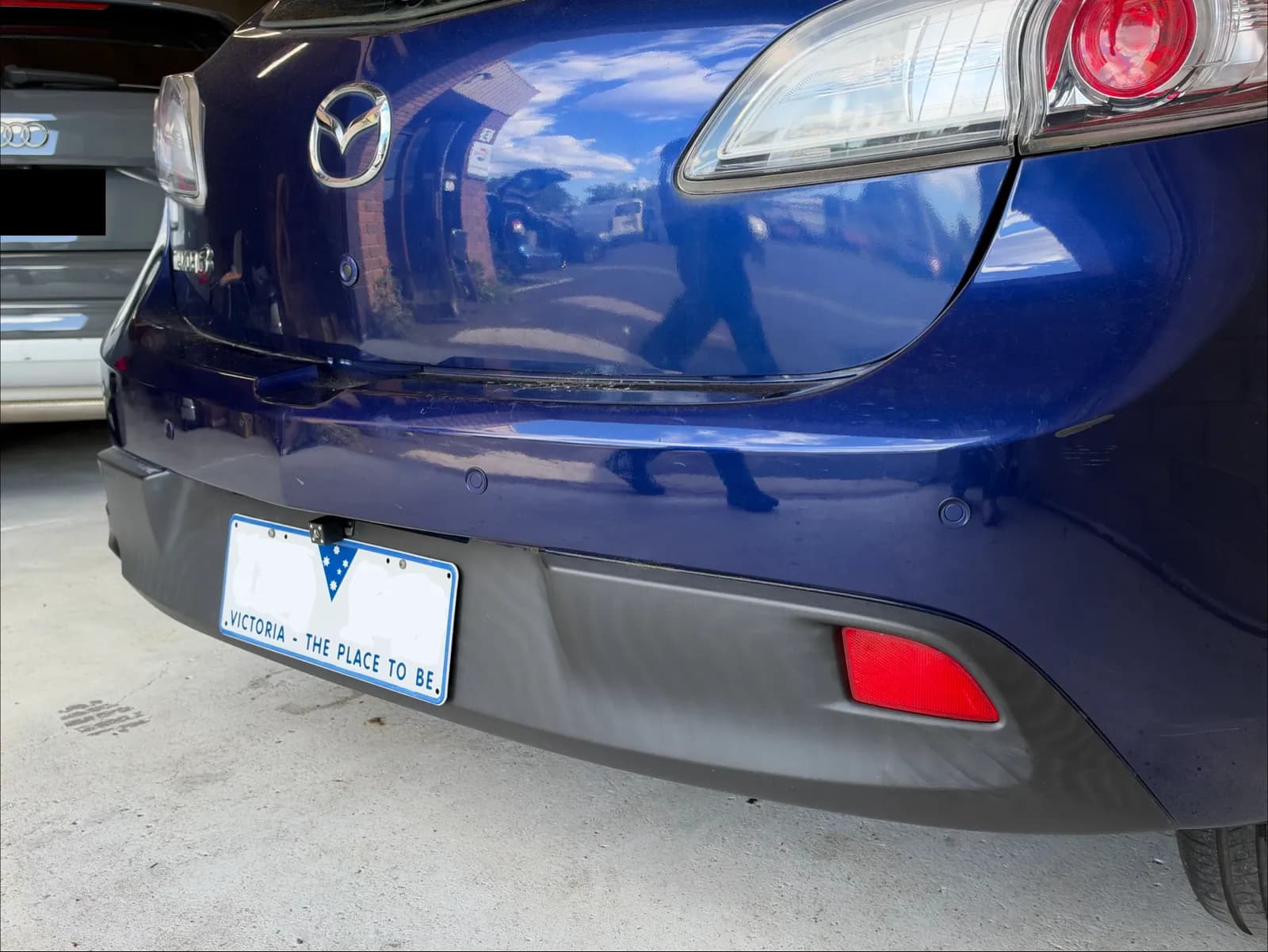 Rear bumper of a 2011 Mazda3 with parking sensors painted to match the blue paint and a reverse camera above the redacted number plate